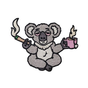 May include: An embroidered patch featuring a grey koala sitting in a cross-legged position. The koala has sleepy eyes, holds a lit brown cigarette with rising smoke in one hand, and a pink mug with steaming liquid in the other hand.