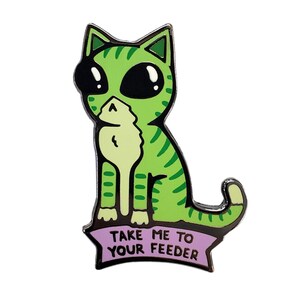May include: Green cat enamel pin with large eyes and a striped pattern. The cat is sitting with its paws tucked under its body. The pin has a purple banner with the text "Take me to your feeder".