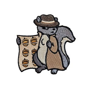 May include: Embroidered patch featuring a cartoon squirrel wearing a fedora and trench coat, holding a scroll with burger illustrations. The squirrel is gray, brown, and beige, with a white background.