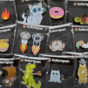 May include: A collection of enamel pins featuring various designs, including a cat, a donut, a lotus flower, a dinosaur, and a ghost. The pins are packaged in clear plastic bags with black backgrounds and the word "kolorsun" printed on them.