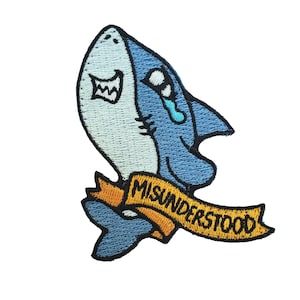 Puede incluir: Un parche bordado con un tiburón de dibujos animados con una lágrima y una pancarta que dice "MISUNDERSTOOD".
