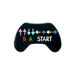 May include: Black enamel pin shaped like a video game controller with colourful directional arrows, B, A, and START buttons.