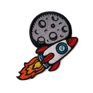 Puede incluir: Un parche bordado que representa un cohete despegando de la superficie de la luna. El cohete es rojo, blanco y azul con llamas naranjas. La luna es gris con cráteres negros.