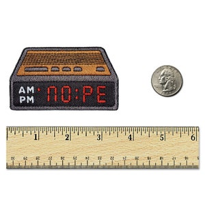 May include: A rectangular embroidered patch shaped like a digital alarm clock with a dark gray border, a tan top section, and a black display area featuring the red text NO:PE. A US quarter and a wooden ruler showing 6 inches are placed nearby.