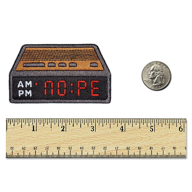 May include: A rectangular embroidered patch shaped like a digital alarm clock with a dark gray border, a tan top section, and a black display area featuring the red text NO:PE. A US quarter and a wooden ruler showing 6 inches are placed nearby.