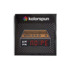 May include: A rectangular embroidered patch featuring a digital alarm clock design with a brown top, black base, and red digital display reading NO:PE. The text AM and PM appears in white on the left. The brand name kolorspun is printed above.