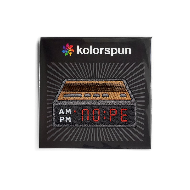May include: A rectangular embroidered patch featuring a digital alarm clock design with a brown top, black base, and red digital display reading NO:PE. The text AM and PM appears in white on the left. The brand name kolorspun is printed above.