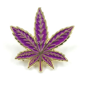 May include: A gold-colored metal pin in the shape of a purple marijuana leaf.