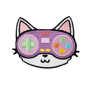 Puede incluir: Parche bordado con un diseño en forma de gato con un mando de videojuego. El mando es morado con una cruz verde, botones coloridos y un contorno rosa. El gato tiene orejas rosas y una cara blanca.