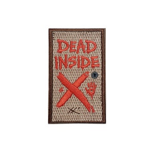 Puede incluir: Un parche bordado marrón con el texto "DEAD INSIDE" en letras rojas. Una X roja con un símbolo de mano y ojo está en el centro del parche.