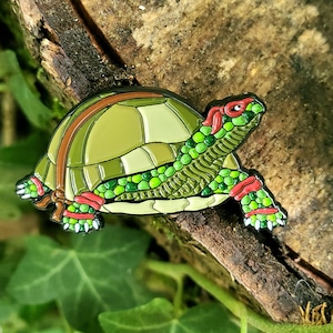 May include: A green and red enamel pin of a turtle with a detailed shell and red accents around the eyes and legs.