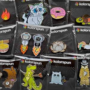 May include: A collection of enamel pins featuring various designs, including a cat, a doughnut, a lotus flower, a dinosaur, and a ghost. The pins are packaged in clear plastic bags with black backgrounds and the word "kolorsun" printed on them.