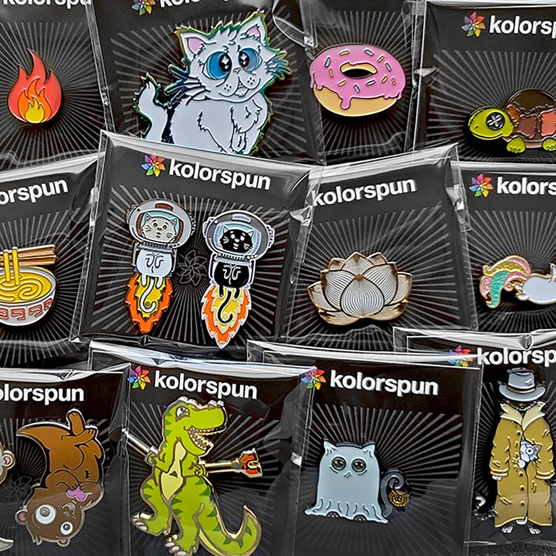 May include: A collection of enamel pins featuring various designs, including a cat, a doughnut, a lotus flower, a dinosaur, and a ghost. The pins are packaged in clear plastic bags with black backgrounds and the word "kolorsun" printed on them.