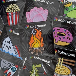 May include: Assortment of embroidered iron-on patches featuring various designs, including a campfire, a donut, a flower, a frog, a rocket ship, a dinosaur, and a bowl of noodles. The patches are all on black backgrounds with a white starburst pattern.