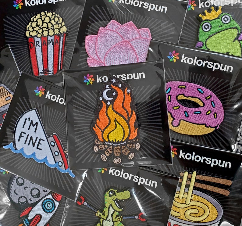 May include: Assortment of embroidered iron-on patches featuring various designs, including a campfire, a donut, a flower, a frog, a rocket ship, a dinosaur, and a bowl of noodles. The patches are all on black backgrounds with a white starburst pattern.