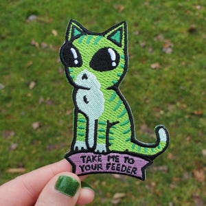 May include: An embroidered patch featuring a green cat with dark alien-like eyes and stripes. The cat has a white chest and sits above a purple banner with the black embroidered text "TAKE ME TO YOUR FEEDER" in a bold, sans-serif font.