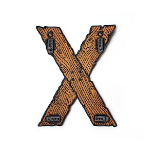 Puede incluir: Un parche bordado marrón en forma de letra X. El parche está diseñado para parecer dos piezas de madera cruzadas una sobre la otra. La madera tiene un patrón de veta y hay detalles de costura negra alrededor de los bordes.