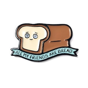 May include: A brown and white enamel pin with a cartoon bread slice with a face and the text "All my friends are bread" on a blue ribbon.