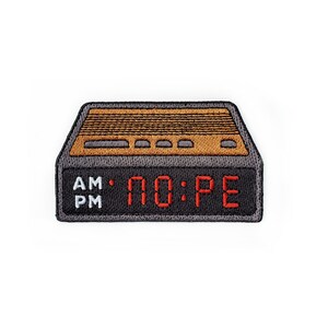 May include: A rectangular embroidered patch shaped like a vintage digital alarm clock. The top section is brown with horizontal lines and buttons. The bottom black section features a red digital display reading NO:PE with AM and PM text.