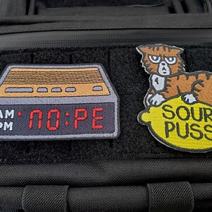 May include: Two embroidered patches attached to a black tactical bag. The left patch is a digital alarm clock displaying the text NO:PE in red. The right patch features a brown tabby cat sitting on a yellow lemon with the text SOUR PUSS below.