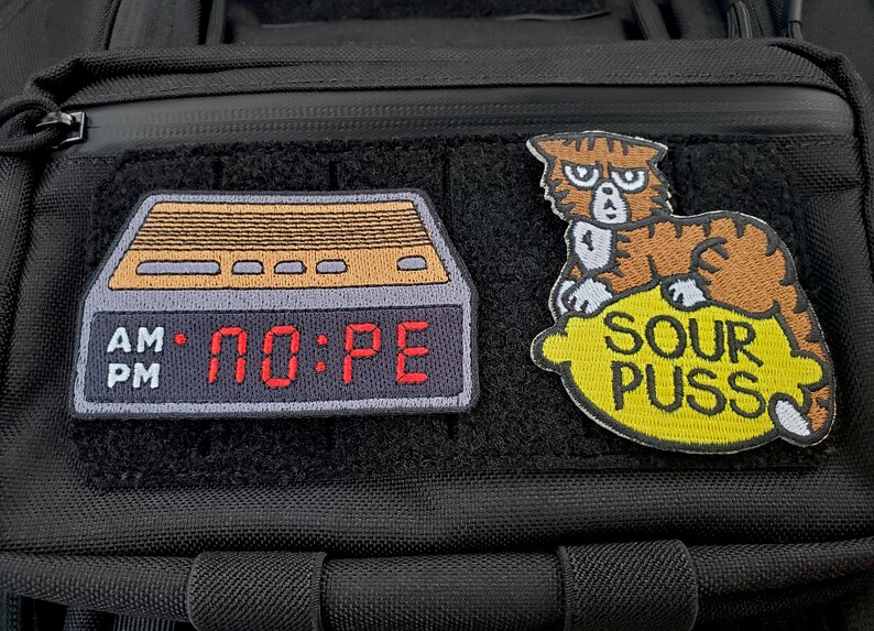 May include: Two embroidered patches attached to a black tactical bag. The left patch is a digital alarm clock displaying the text NO:PE in red. The right patch features a brown tabby cat sitting on a yellow lemon with the text SOUR PUSS below.