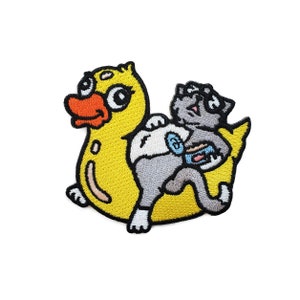 May include: Embroidered patch featuring a yellow duck with an orange beak and a gray cat. The cat is holding a small object. The patch has a black border and is designed for application on clothing or accessories.