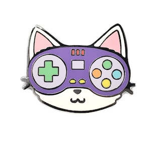 May include: Purple and white enamel pin of a cat wearing a purple video game controller over its eyes. The controller has green, yellow, orange, and blue buttons.