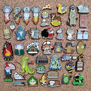 May include: A collection of assorted enamel pins featuring various cartoon characters, animals, food items, and humorous text designs. The pins include illustrations of cats, raccoons, a dinosaur, a gnome, and objects like a slice of pizza and a clock.