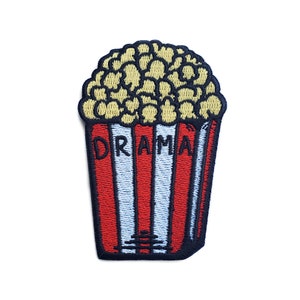 May include: Embroidered iron-on patch featuring a red and white striped popcorn bucket with the word "DRAMA" written on it. The popcorn bucket is filled with yellow popcorn.