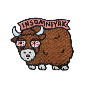 Puede incluir: Parche bordado con un yak marrón con ojos rojos y blancos y una pancarta rosa encima que dice "Insomniyak".