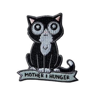 Puede incluir: Parche bordado con un gato de dibujos animados con ojos grandes, pelaje negro y un babero blanco. El gato está sentado sobre una pancarta que dice "MOTHER I HUNGER". El parche tiene un fondo blanco.