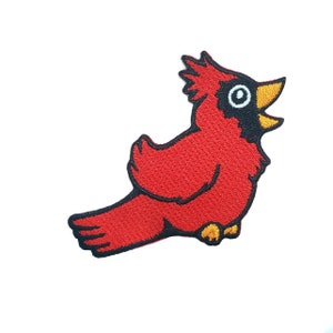 Puede incluir: Un pájaro cardenal rojo con ojos negros y pico amarillo, cantando con la boca abierta. El pájaro está bordado sobre un fondo blanco.
