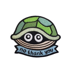 May include: Embroidered patch featuring a cartoon turtle with its head and eyes peeking out from its shell. The turtle is green and blue with a beige shell. The patch has a blue banner with the text "no thank you".