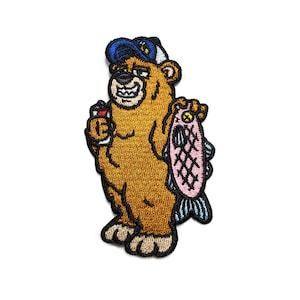 Puede incluir: Parche bordado de un oso marrón que lleva una gorra de béisbol azul, sosteniendo una lata y un pez rosa. El oso tiene una sonrisa de dientes y lleva una gorra de béisbol azul con visera blanca.