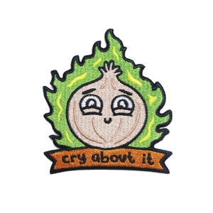 May include: An embroidered patch featuring a cartoon onion with a green flame-like head and a smiling face. The text "cry about it" is written on a brown banner below the onion.