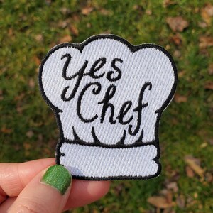 May include: A white embroidered patch shaped like a chef hat with a black stitched border. The text "yes Chef" is embroidered in black cursive lettering across the center of the hat. The patch features a textured fabric finish throughout.