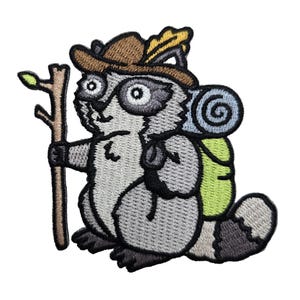 May include: Embroidered patch of a cartoon raccoon dressed as a hiker. The raccoon wears a brown hat with a yellow feather, carries a wooden staff, and has a green backpack with a rolled-up blue blanket. The raccoon is gray, black, and white.