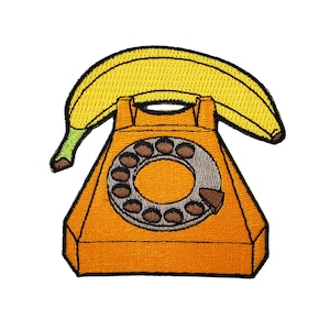May include: A vintage-style orange rotary phone with a yellow banana forming the handset. The phone has a round dial with brown numbers and a gray rotary dial.