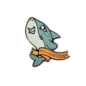 May include: A blue and white enamel pin of a cartoon shark with a tear coming from its eye. The shark is wearing a yellow banner that says "MISUNDERSTOOD".