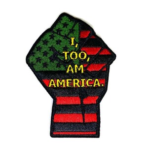 May include: A black and green fist raised in the air with a red, white, and blue American flag design. The text "I, TOO, AM AMERICA." is embroidered in yellow on the fist.