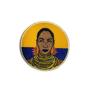 May include: A round embroidered patch featuring a portrait of a woman with dark skin, wearing large gold hoop earrings and a gold necklace. The background is a yellow circle with a blue and orange horizon line.