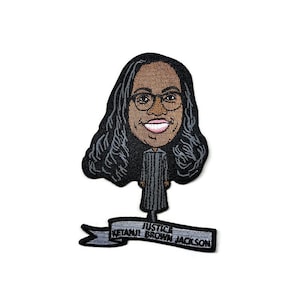 May include: Embroidered patch featuring a cartoon illustration of Justice Ketanji Brown Jackson with black hair, glasses, and a judicial robe. The patch includes the text "JUSTICE KETANJI BROWN JACKSON" on a banner below the figure.