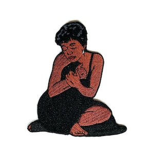 May include: Embroidered patch of a woman in a black dress, sitting with her arms around a small child. The woman has short black hair and brown skin. The patch is made of felt and has a stitched outline.