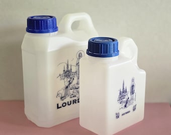 Puede incluir: Dos jarras de agua de plástico blanco con tapas de rosca azules. Cada jarra tiene un asa y una ilustración azul de una iglesia y texto. La jarra más grande dice "LOURDES" y la más pequeña dice "LOCURDES" y "AVE MARIA".