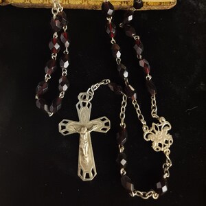 Op de afbeelding: Een zilveren rozenkrans met een kruisbeeld en donkerrode glazen kralen.
