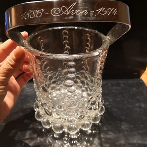Op de afbeelding: Een glazen ijs emmer met een zilveren metalen handvat. Het handvat is gegraveerd met "1886 - Avon - 1944".