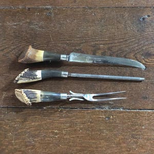 May include: A three-piece carving set featuring a knife, a sharpening steel, and a two-pronged fork. Each utensil has a handle made of textured, brown and cream-colored antler with a silver-toned metal bolster connecting to the steel blade.