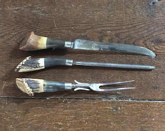 May include: A three-piece carving set featuring a knife, a sharpening steel, and a two-pronged fork. Each utensil has a handle made of textured, brown and cream-colored antler with a silver-toned metal bolster connecting to the steel blade.