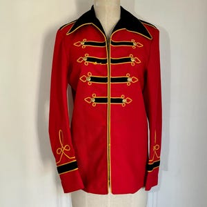 May include: A bright red long-sleeve jacket featuring a black collar and black horizontal bands across the front. The jacket is accented with gold-colored piping, decorative gold buttons, and gold embroidery details on the cuffs and front.