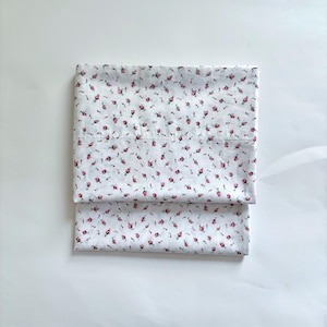 May include: Two folded white pillowcases with a repeating pattern of small red roses and green leaves. The pillowcases are made of a soft, lightweight fabric, perfect for a bedroom.